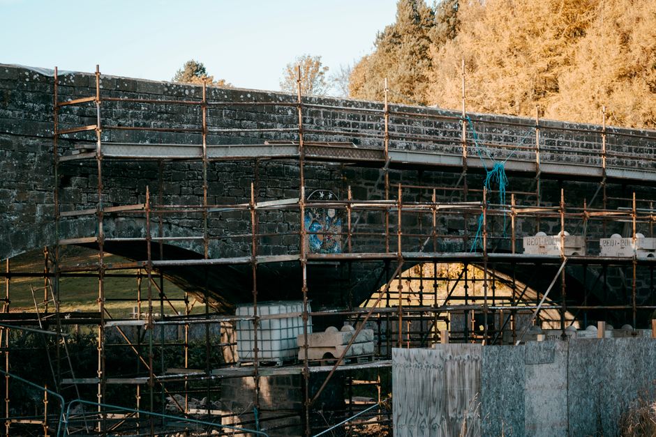 Stone bridge undergoing restoration with autumn trees in the background.