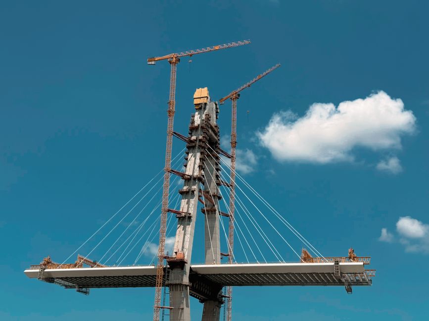 Dynamic view of a cable-stayed bridge under construction with cranes against a clear blue sky.